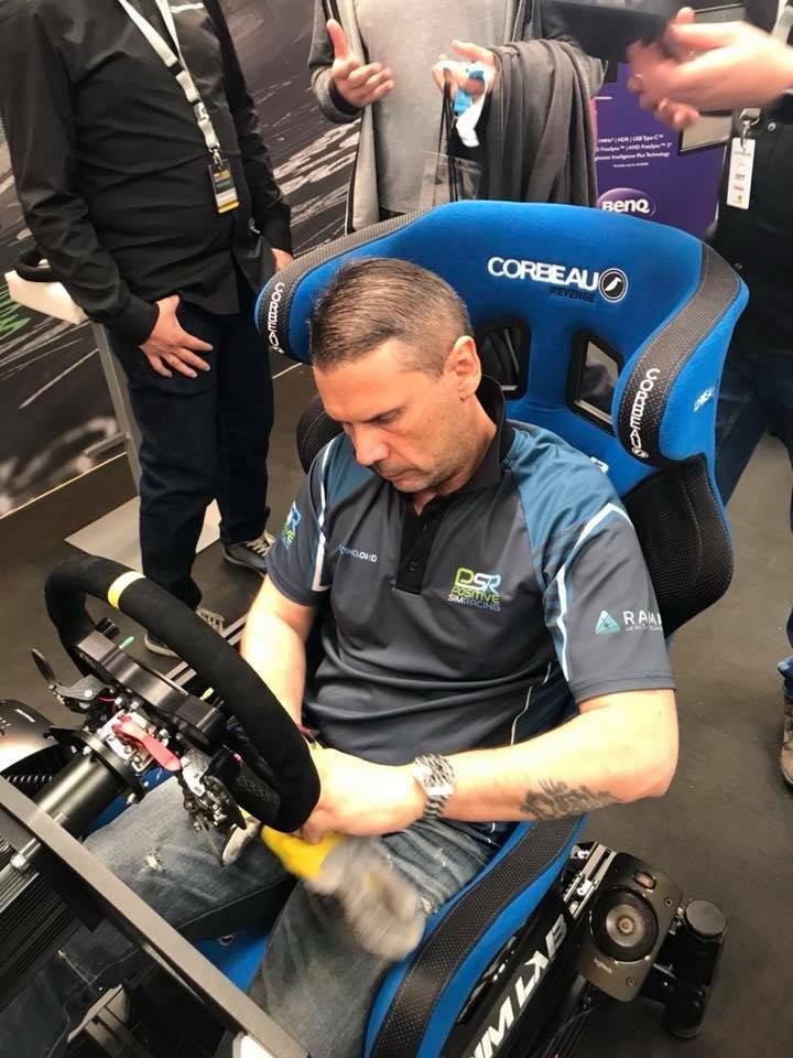 Man seated in a racing simulator chair, adjusting the steering wheel with a tool at an event.