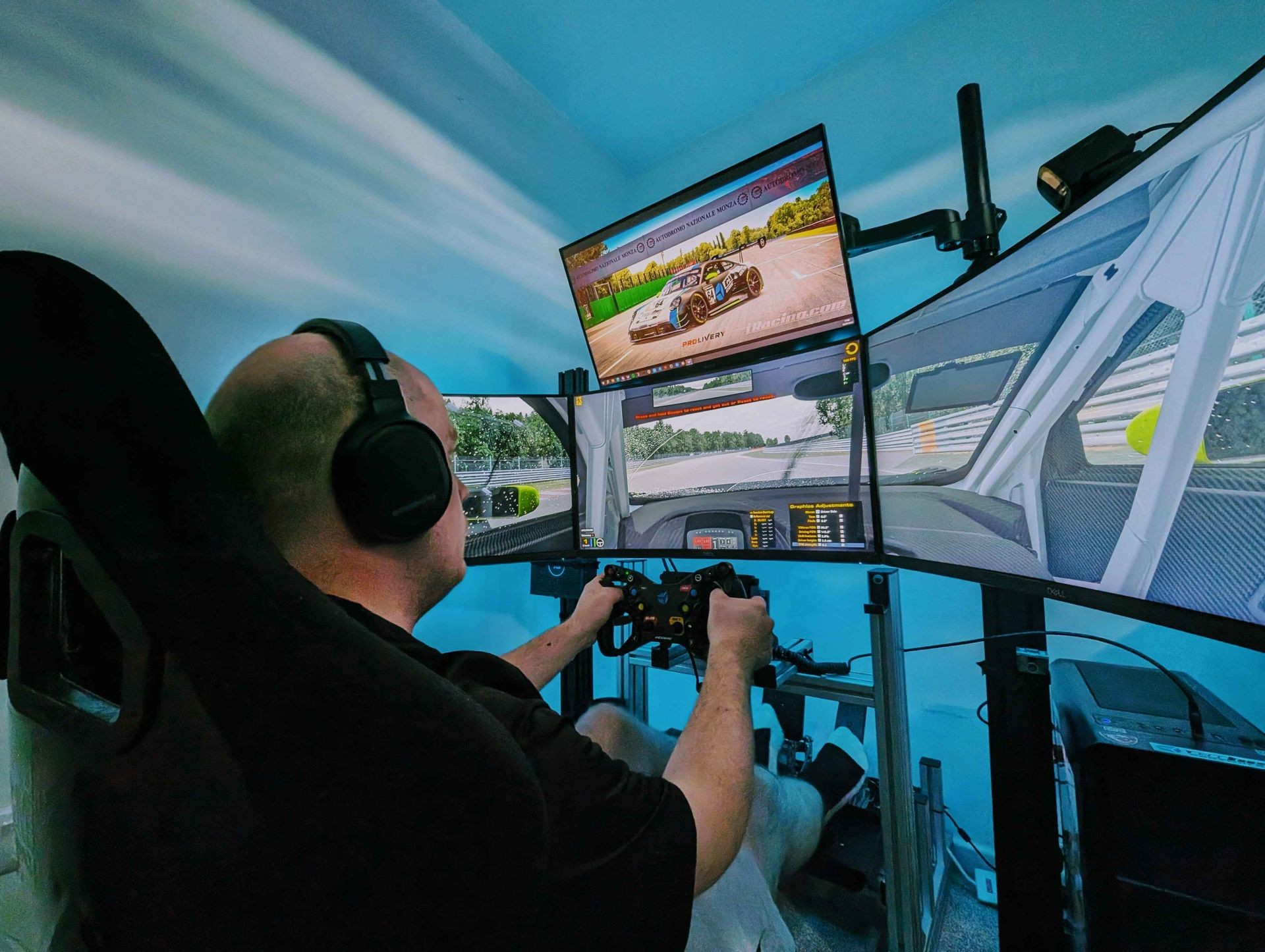 Person using a racing simulator setup with multiple monitors and gaming controls in a blue-lit room.